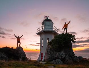 Two people standing on rocks in front of the Baring Head Lighthouse at sunset.