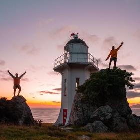 Two people standing on rocks in front of the Baring Head Lighthouse at sunset.