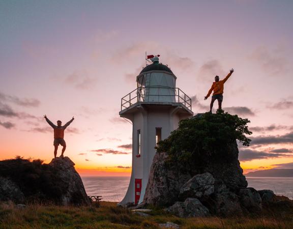 Two people standing on rocks in front of the Baring Head Lighthouse at sunset.