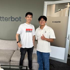 Two smiling participants in the Summer of Engineering are standing in a room, smiling in front of a sign that says 'Clutterbot'. 