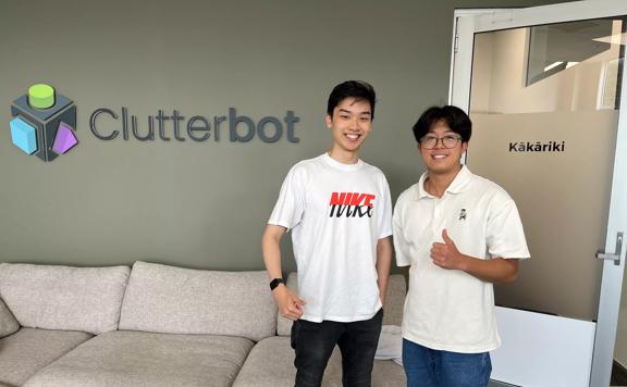Two smiling participants in the Summer of Engineering are standing in a room, smiling in front of a sign that says 'Clutterbot'. 