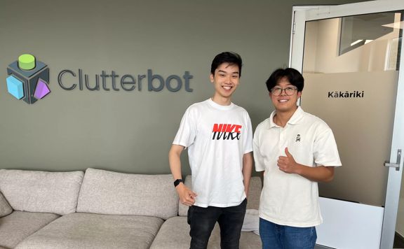 Two smiling participants in the Summer of Engineering are standing in a room, smiling in front of a sign that says 'Clutterbot'.