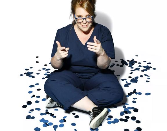 Georgie Carroll sits cross-legged on the floor in blue scrubs with blue confetti surrounding her.