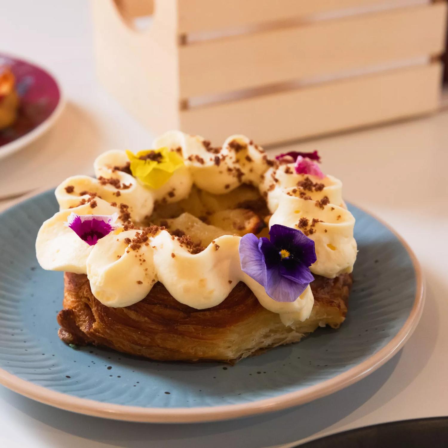 A circular pastry dessert with fruit filling, white icing and yellow, pink and purple flowers on a pastel-blue plate at Le Ciel Bakery in Te Aro, Wellington. 