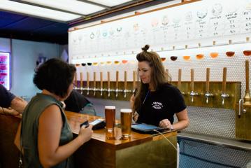 Beer taps line the tiled wall of the Garage Project Taproom while a person is served two beers by an employee.