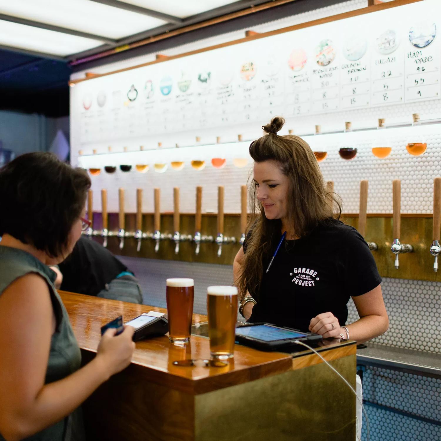 Beer taps line the tiled wall of the Garage Project Taproom while a person is served two beers by an employee.