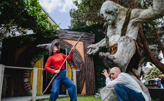 Two visitors posing with a giant troll sculpture at Wētā Workshop, with one holding a staff outside a hobbit-style building surrounded by trees and greenery.