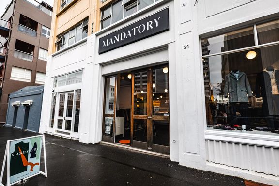 The exterior of Mandatory, with wooden doors and white painting outside. Mannequins are seen in the windows wearing their clothing.