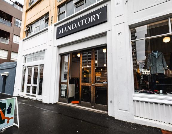 The exterior of Mandatory, with wooden doors and white painting outside. Mannequins are seen in the windows wearing their clothing.