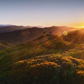 The sun rises over bushy hills. Two people stand next to a trig on one of the hills.