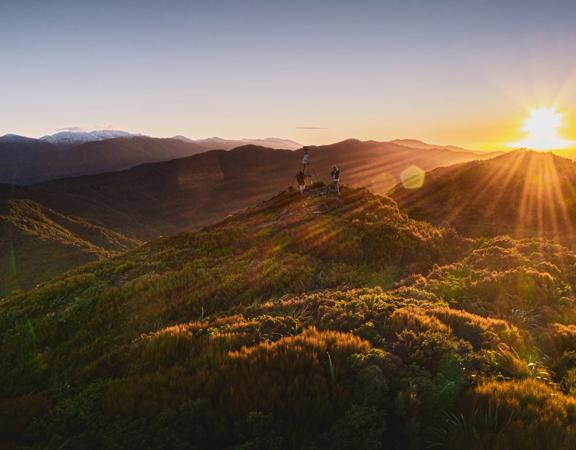 The sun rises over bushy hills. Two people stand next to a trig on one of the hills.