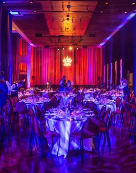 A room inside the Michael Fowler Centre where a ball is being held, purple, blue, and red lights shinedown on tables set up with waiters walking around.