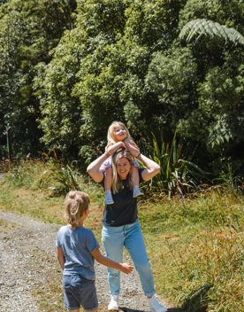 An adult carries a small child on their shoulders while walking along a nature trail with another kid beside them.