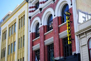 The exterior of The Intrepid Hotel in Te Aro, Wellington. It is a red-brick building with white accents, large windows and a vertical sign displaying the hotel's name.