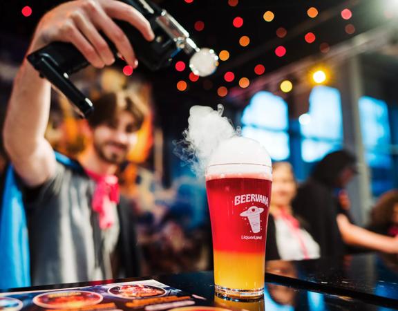 A person holds a tool applying smoke to the top of a pint of red and yellow beer at Beervana 2023, a beer and food festival in Wellington New Zealand.