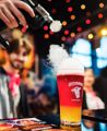 A person holds a tool applying smoke to the top of a pint of red and yellow beer at Beervana 2023, a beer and food festival in Wellington New Zealand.
