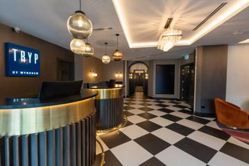 The lobby of TRYP by Wyndham Wellington hotel. There are grey and white walls, black and white tiled floors, two concierge desks and decorative light fixtures on the ceiling.