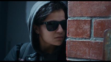 A screen grab from Millie Lies Low, of a person with a hoodie and glasses on spies around a brick wall.