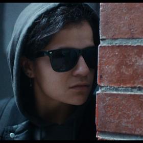 A screen grab from Millie Lies Low, of a person with a hoodie and glasses on spies around a brick wall.