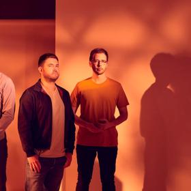 Image of all three members of GoGo Penguin standing against a wall. Shadowy orange lighting is used. 