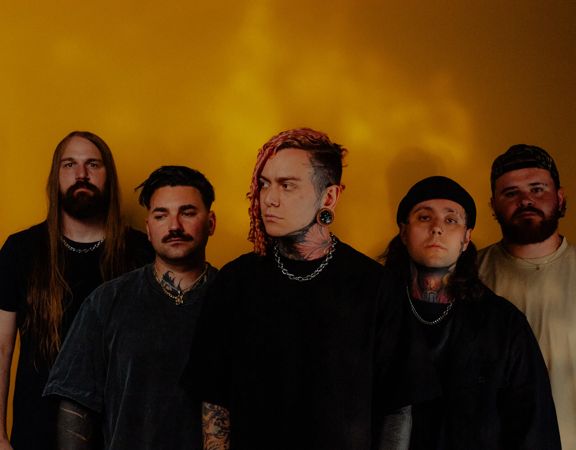 Five band members of Lorna Shore stand in front of a yellow-orange wall.