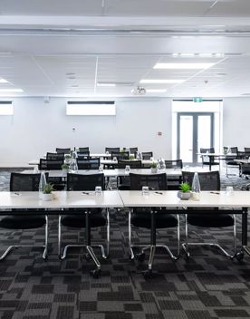 Conference/event room at NZCIS. White tables are set up with four chairs behind each and glasses and house plants sit on the tables. The ceiling and walls are white with grey carpet.