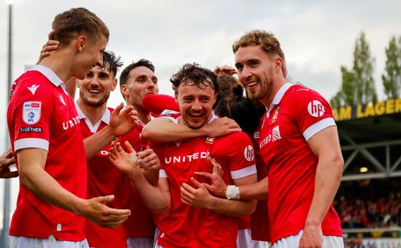 Players from Wrexham AFC huddle together in celebration, they wear read and white shirts.