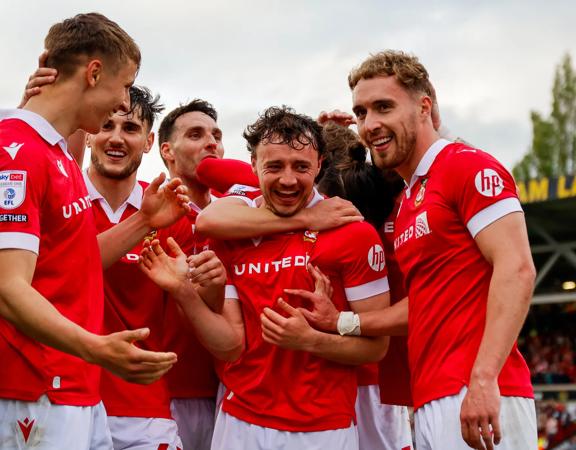 Players from Wrexham AFC huddle together in celebration, they wear read and white shirts.