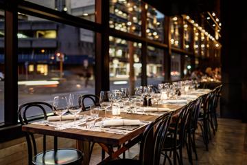 A long rectangular table is set up for a fancy dinner for twelve people in a dimly lit space with high ceilings, large windows and a contemporary industrial vibe.