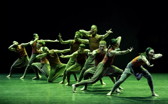 Members of the Akram Khan Company on stage posing with arms outstretched. The background is black but the floor and the performers are bathed in green light.