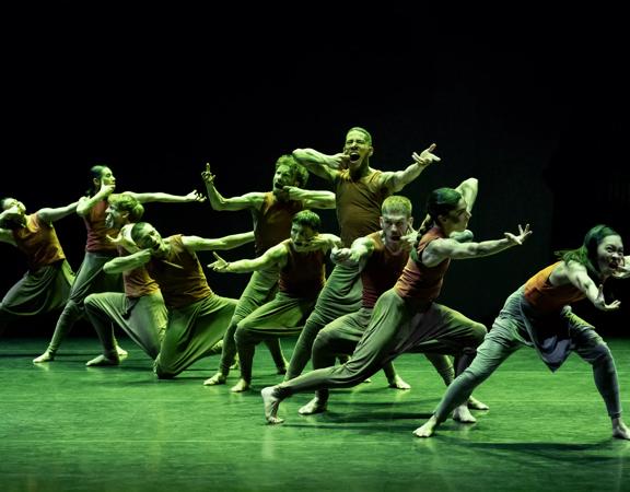 Members of the Akram Khan Company on stage posing with arms outstretched. The background is black but the floor and the performers are bathed in green light.