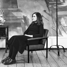 Alison Moyet wears all black and sits in an armchair with a blank stare in a room with hardwood floors and a few antiques scattered around.