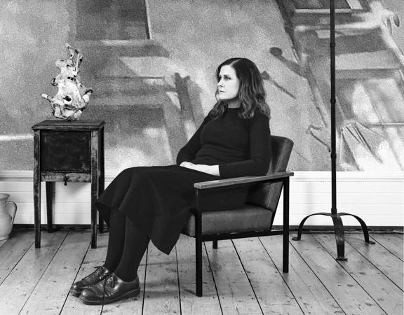 Alison Moyet wears all black and sits in an armchair with a blank stare in a room with hardwood floors and a few antiques scattered around. 