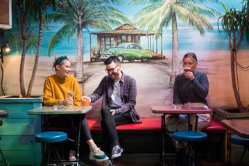 Three people sit on a bench seat against a wall, drinking cocktails. The wall is painted with palm trees, a cabana, and a 50s-style car.