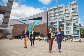 Two pairs of conference attendees with reusable takeaway coffees walk along the promenade in front of the Museum of New Zealand Te Papa Tongarewa on a sunny day in Wellington.