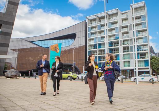 Two pairs of conference attendees with reusable takeaway coffees walk along the promenade in front of the Museum of New Zealand Te Papa Tongarewa on a sunny day in Wellington.