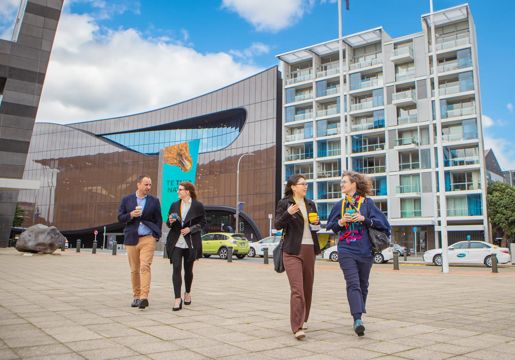 Two pairs of conference attendees with reusable takeaway coffees walk along the promenade in front of the Museum of New Zealand Te Papa Tongarewa on a sunny day in Wellington.