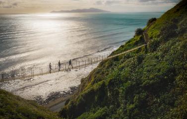 3 people walking along a swing bridge between 2 hillsides on the Escarpment track above the Kāpiti coast.