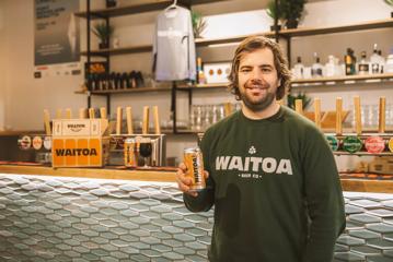 A person in a green shirt stands by the bar and holds a tall can of Wiatoa's ' Earlybird stout in their right hand.