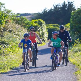 Two adults and two children cycling along a wide, flat path in Queen Elizabeth Park, Kāpiti Coast.