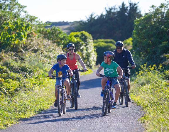 Two adults and two children cycling along a wide, flat path in Queen Elizabeth Park, Kāpiti Coast.