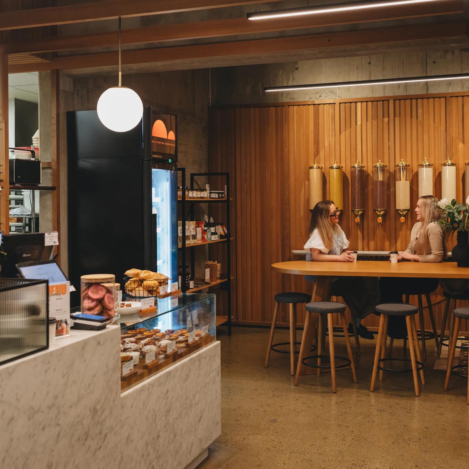 Two people sit at a large table enjoying coffee at Wellington Sourdough. The cabinet is stacked with baked goods.