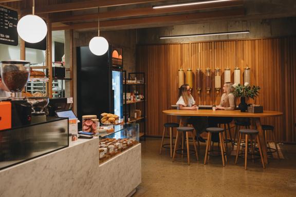 Two people sit at a large table enjoying coffee at Wellington Sourdough. The cabinet is stacked with baked goods.