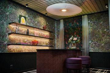 A dimly-lit lounge space with floral wall paper, a paper chandelier and a high-table with two round purple stools.