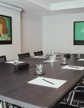 The Mowbray Room, a conference room in the Bolton Hotel in Wellington is set up with four large tables pushed together, eight chairs with a water glass, notepad and pen at each seat, light grey walls and two mounted TVs.