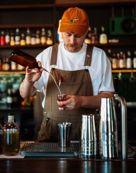 A bartender wearing a white teeshirt, brown overalls, and an orange cap mixes a cocktail behind the bar at Regent.