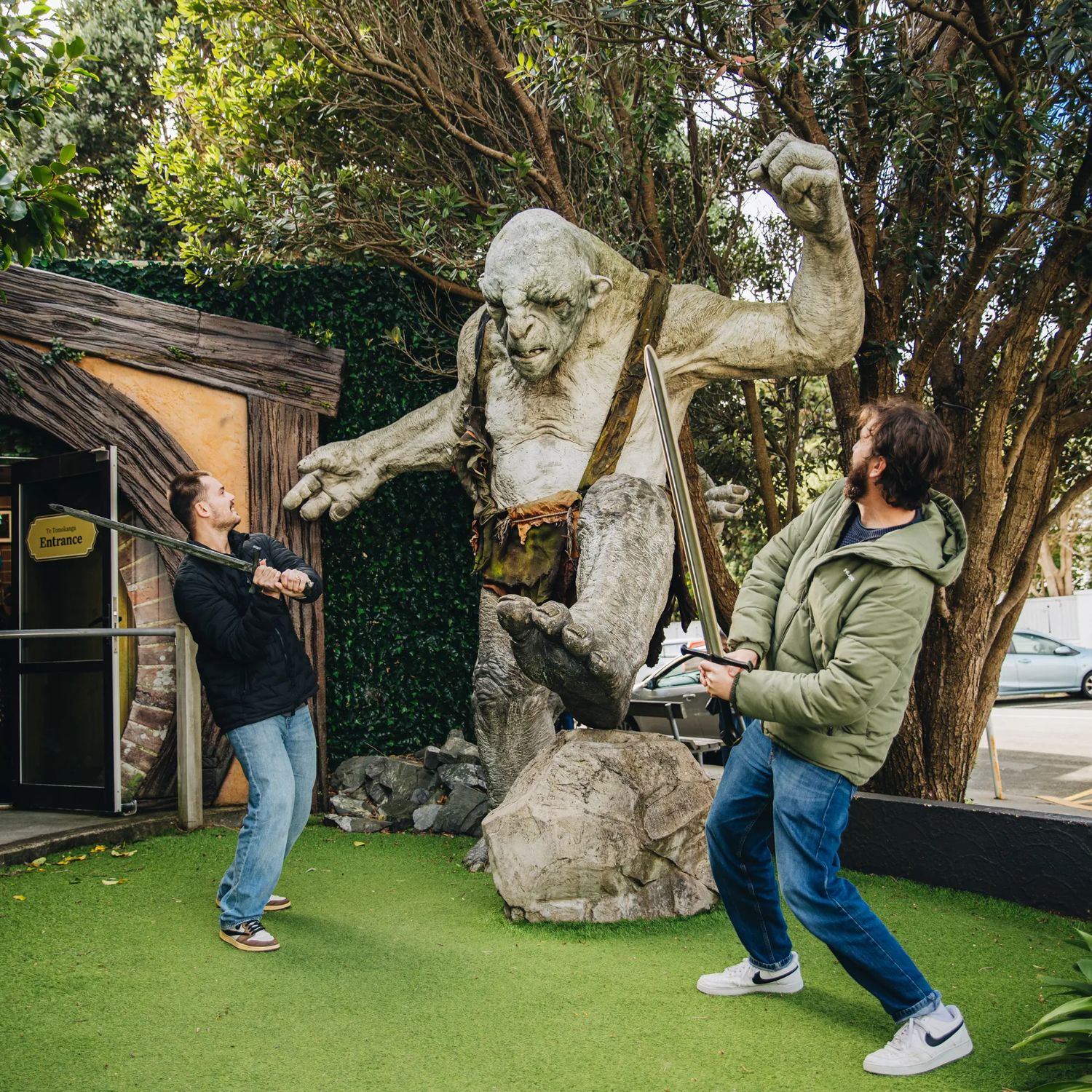 Two visitors pose with a giant troll sculpture at Wētā Workshop, with one holding a staff outside a hobbit-style building surrounded by trees and greenery.
