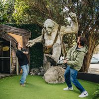 Two visitors pose with a giant troll sculpture at Wētā Workshop, with one holding a staff outside a hobbit-style building surrounded by trees and greenery.