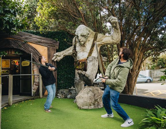 Two visitors pose with a giant troll sculpture at Wētā Workshop, with one holding a staff outside a hobbit-style building surrounded by trees and greenery.