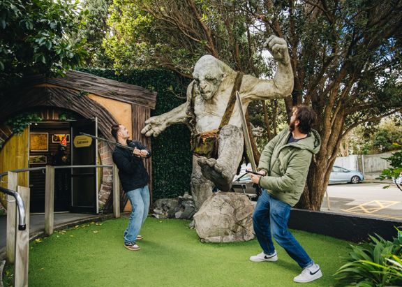 Two visitors pose with a giant troll sculpture at Wētā Workshop, with one holding a staff outside a hobbit-style building surrounded by trees and greenery.